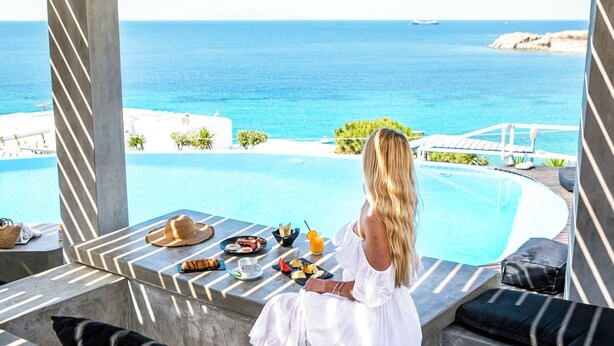 A person sitting by a pool with a view of the sea, a dining table with food and drinks, and lounge chairs. - Apartment with Pool & Shared Infinity Pool, Sea View,  Pool Bar, Gym (C965+8J Mykonos, Řecko)