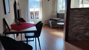 Un área de comedor con una mesa, sillas y un mantel individual rojo. Se ve un sofá y una pequeña mesa redonda al fondo.
