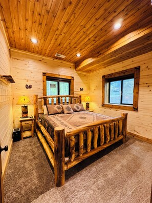 Ein Schlafzimmer einer Holzhütte mit einem großen Bett, zwei Fenstern und einem kleinen Nachttisch mit Lampen.