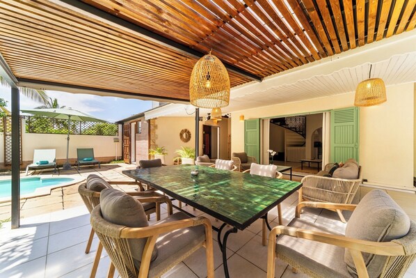An outdoor dining area with a glass table and wicker chairs, a pool, and a covered patio with wooden beams and hanging lights.