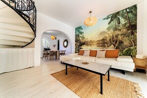 Un salon avec un canapé blanc, une table basse et une grande fresque murale représentant un paysage tropical.