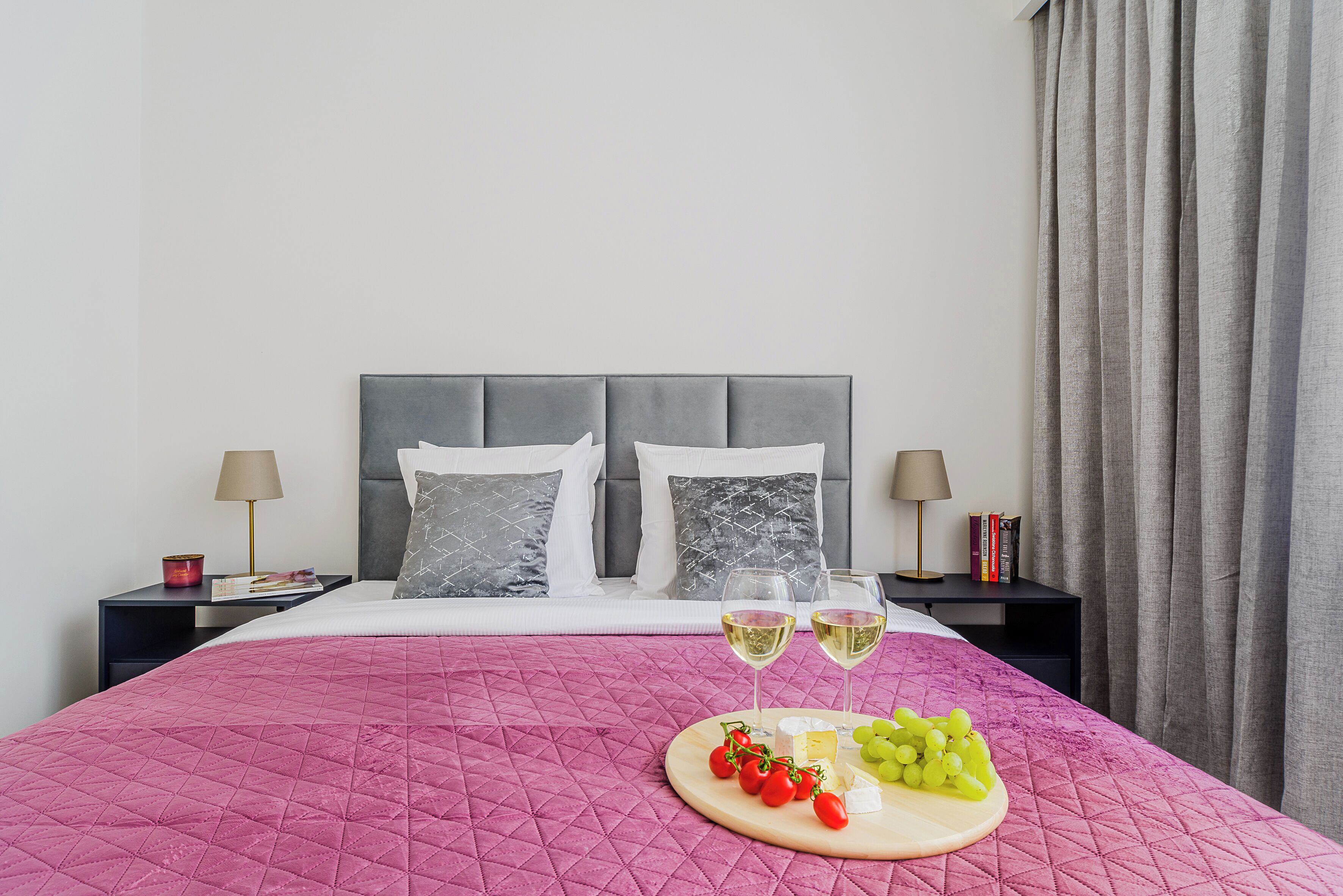 A neatly made bed with a pink quilt, two bedside tables with lamps, a tray of cheese and grapes, and two glasses of white wine.