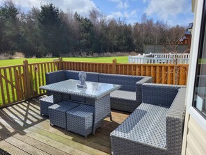A covered outdoor seating area with wicker furniture and a glass-top table, surrounded by a wooden deck and a wooden fence.