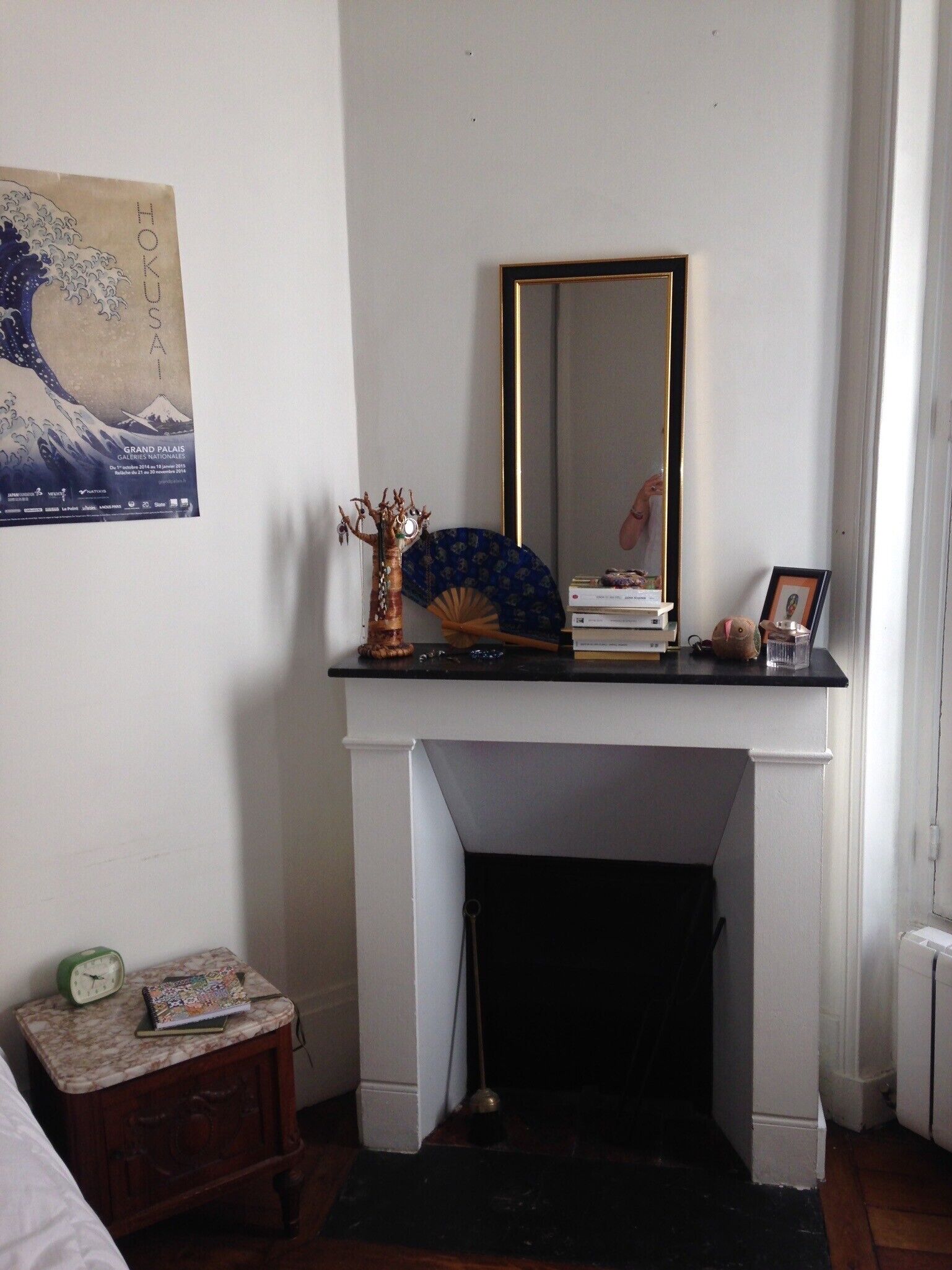 A room with a white fireplace, a mirror above it, a small table with a clock and a book, and a decorative fan.