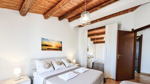 A bedroom with a wooden ceiling, a bed with white bedding, a bedside table with a lamp, and a painting of a sunset on the wall. - MareLuna by Interhome (Fontane Bianche)