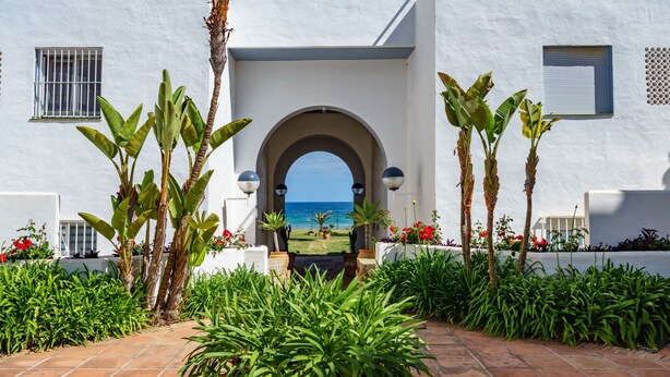 A white building with a tiled entrance, a view of the sea through an archway, and tropical plants. - Apartment Ayla by Interhome (Casares)