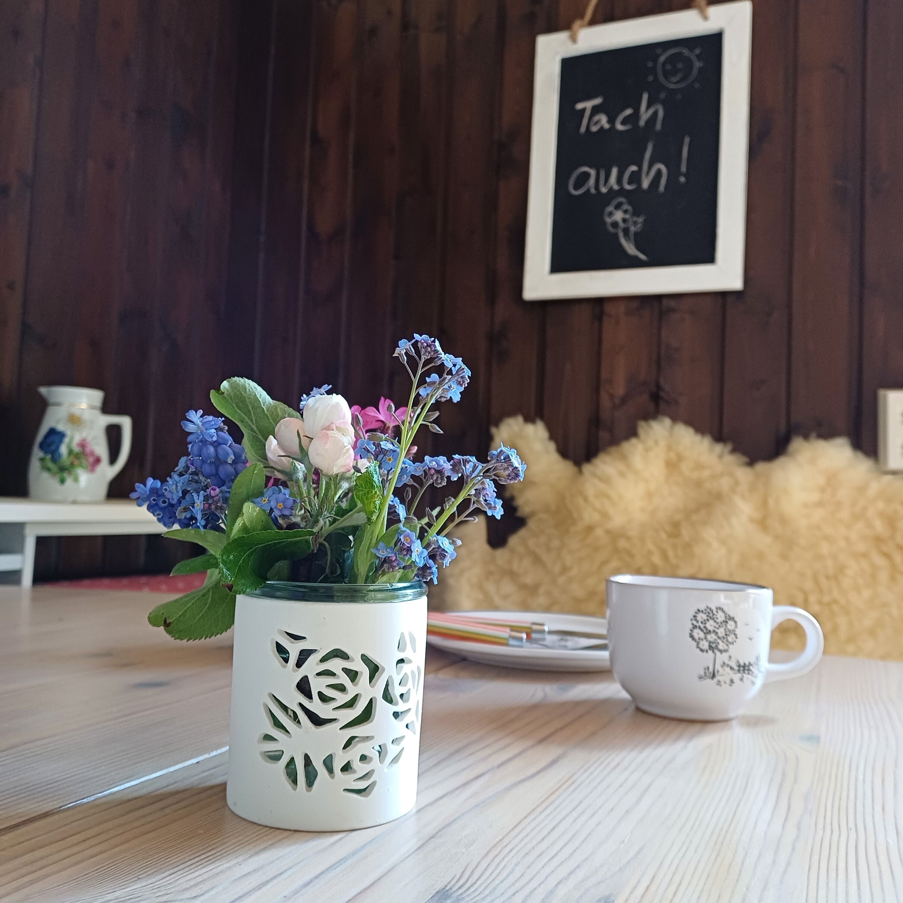 Puisesta pöydästä löytyy koristeellinen kukilla täytetty muki, valkoinen kukkapainokuosin omaava muki sekä taustalla oleva kyltti, jossa on tekstiä.