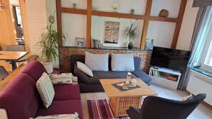 A cozy living room with a sofa, armchair, and coffee table. There is a TV, bookshelf, and a window with curtains.