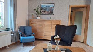 A modern living room with a blue armchair, a black armchair, a wooden cabinet, and a dining table with a decorative hourglass.