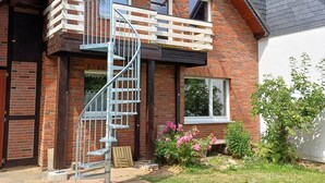 A two-story house with a wooden balcony, a spiral staircase, and a garden with plants and flowers.