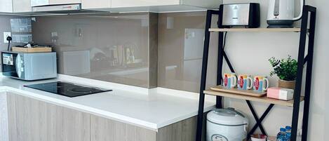 Une cuisine moderne dotée d’un îlot central, d’un four encastré et d’une machine à café. On y trouve une étagère avec des tasses et une bouilloire, ainsi qu’une poubelle.