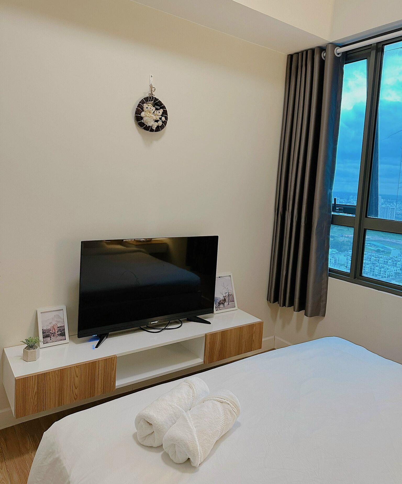 A hotel room with a flat-screen TV on a wooden TV stand, a bed with white bedding, a small potted plant, and a view of the city through a window with dark curtains.