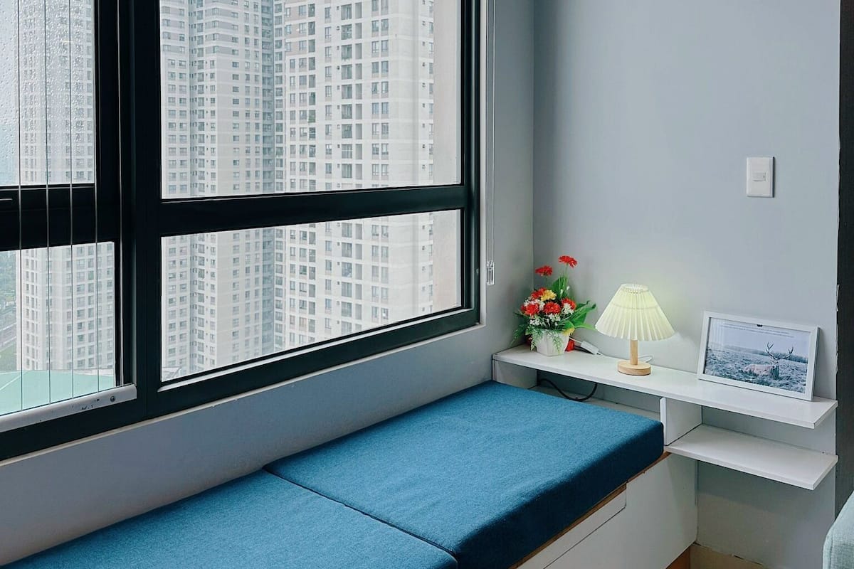 Une pièce avec une grande fenêtre, un banc bleu avec un coussin, une petite table avec une lampe et un cadre photo.