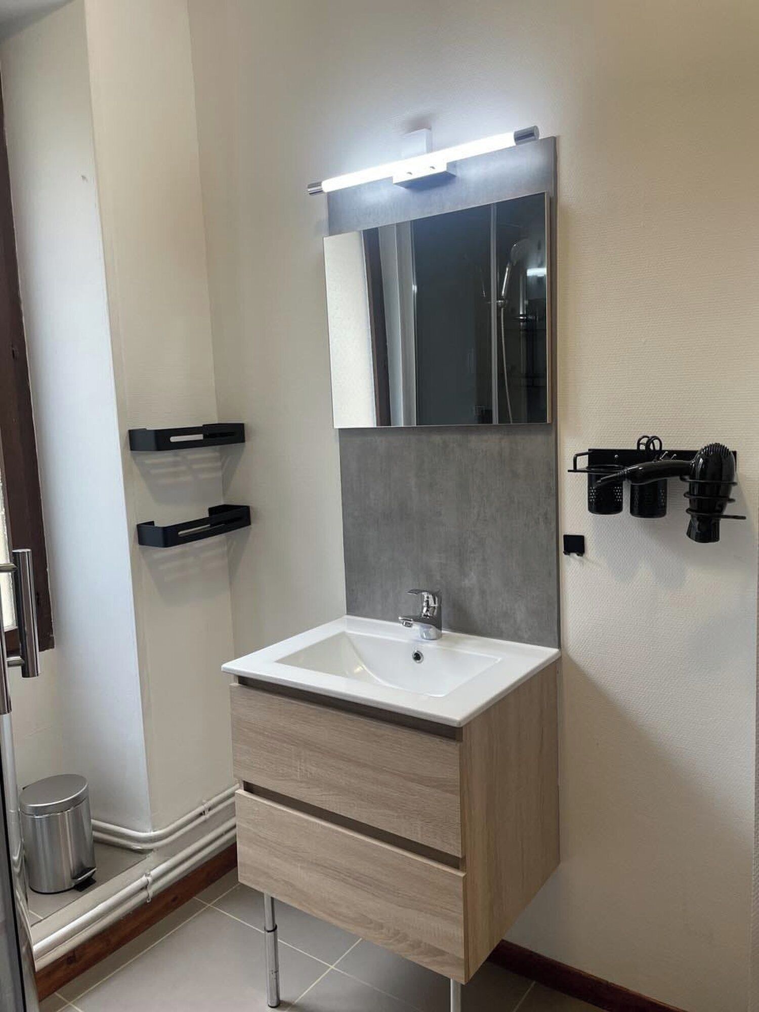 Een moderne badkamer met een wit wastafelmeubel, een spiegel en een douche. Er zitten zwarte handdoekhaken aan de muur en een prullenbak naast het wastafelmeubel.
