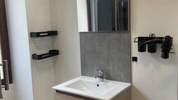 Een moderne badkamer met een wit wastafelmeubel, een spiegel en een douche. Er zitten zwarte handdoekhaken aan de muur en een prullenbak naast het wastafelmeubel.