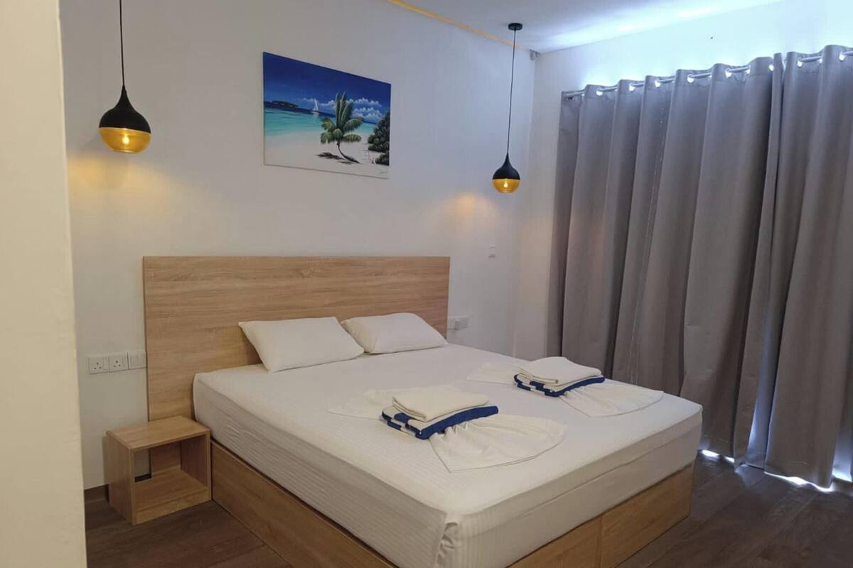 A neatly made bed with white linens and two pillows, a wooden headboard, a small bedside table, and two pendant lights.