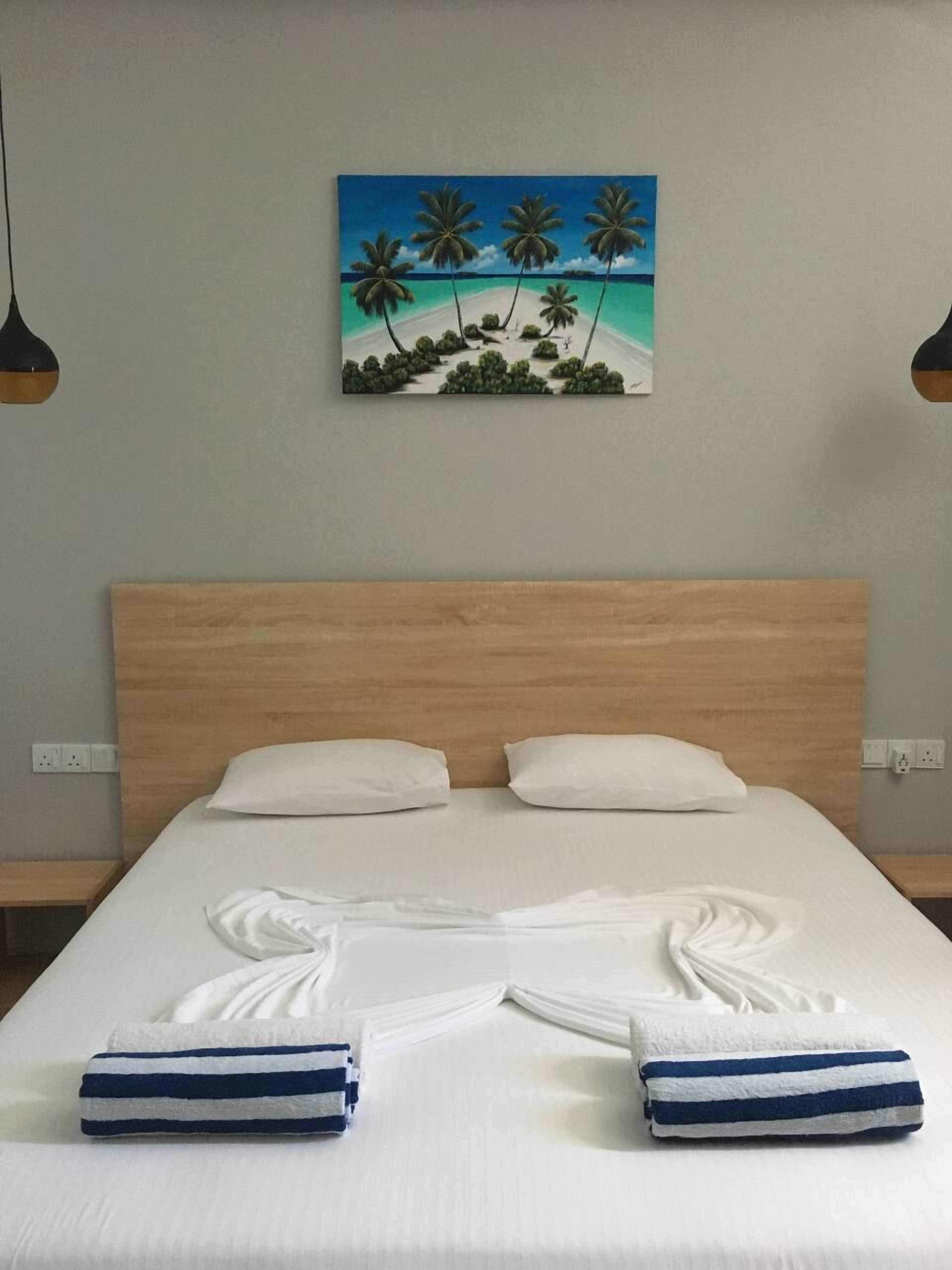 A hotel room with a wooden headboard, two white pillows, a neatly made bed with a white and blue striped towel, and a painting of a tropical beach on the wall.