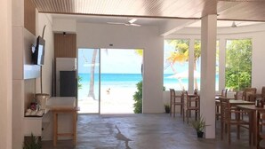 A spacious room with a view of the beach, a dining area with wooden furniture, and a ceiling with wooden slats. - The Sunset Villa (Dhiffushi)