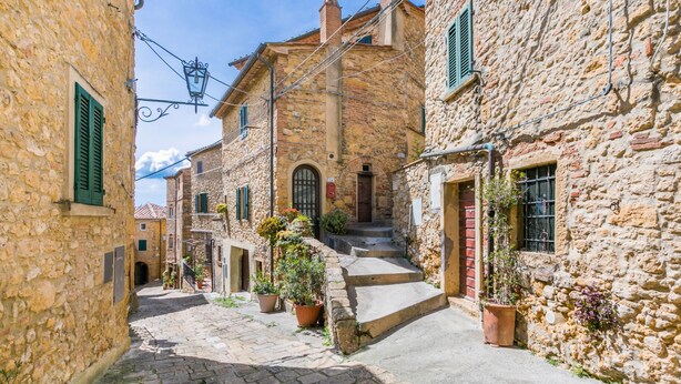 Una estrecha calle empedrada bordeada por edificios de piedra con puertas arqueadas y contraventanas verdes.