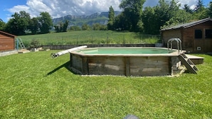 Une piscine en bois avec un escalier, entourée d’une pelouse verdoyante, et en arrière-plan, un chalet en bois et des montagnes.