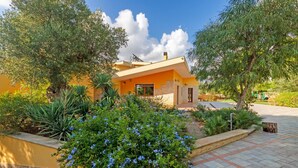 Une maison moderne à la façade jaune, entourée d’une végétation luxuriante et de fleurs bleues.