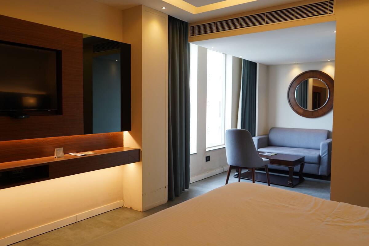 A hotel room with a large bed, a desk with a flat-screen TV, a seating area with a sofa and armchair, and a large window with curtains.