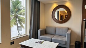 A modern hotel room with a grey sofa, a marble coffee table, and a large window with a view of palm trees. - The Sea View (Mangaluru)