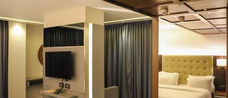 A hotel room with a bed, a desk with a flat-screen TV, a small seating area, and a large window with curtains.
