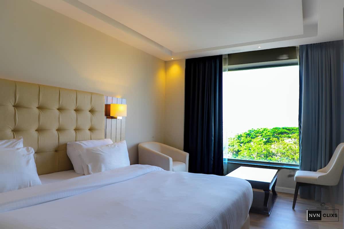 A hotel room with a large bed, a small table, two chairs, and a view of greenery through a window.