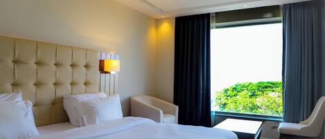 A hotel room with a large bed, a small table, two chairs, and a view of greenery through a window.