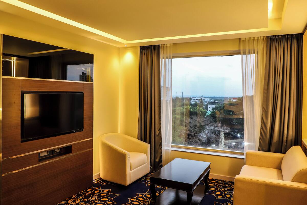 A hotel room with a large TV, two sofas, a coffee table, and a view of a cityscape through a large window.