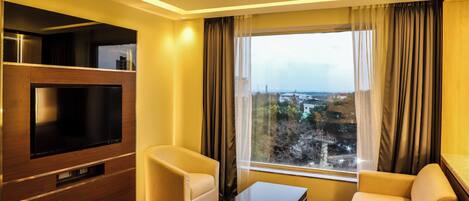 Habitación de hotel con un televisor grande, dos sofás, una mesa de centro y vistas a un paisaje urbano a través de un ventanal amplio.