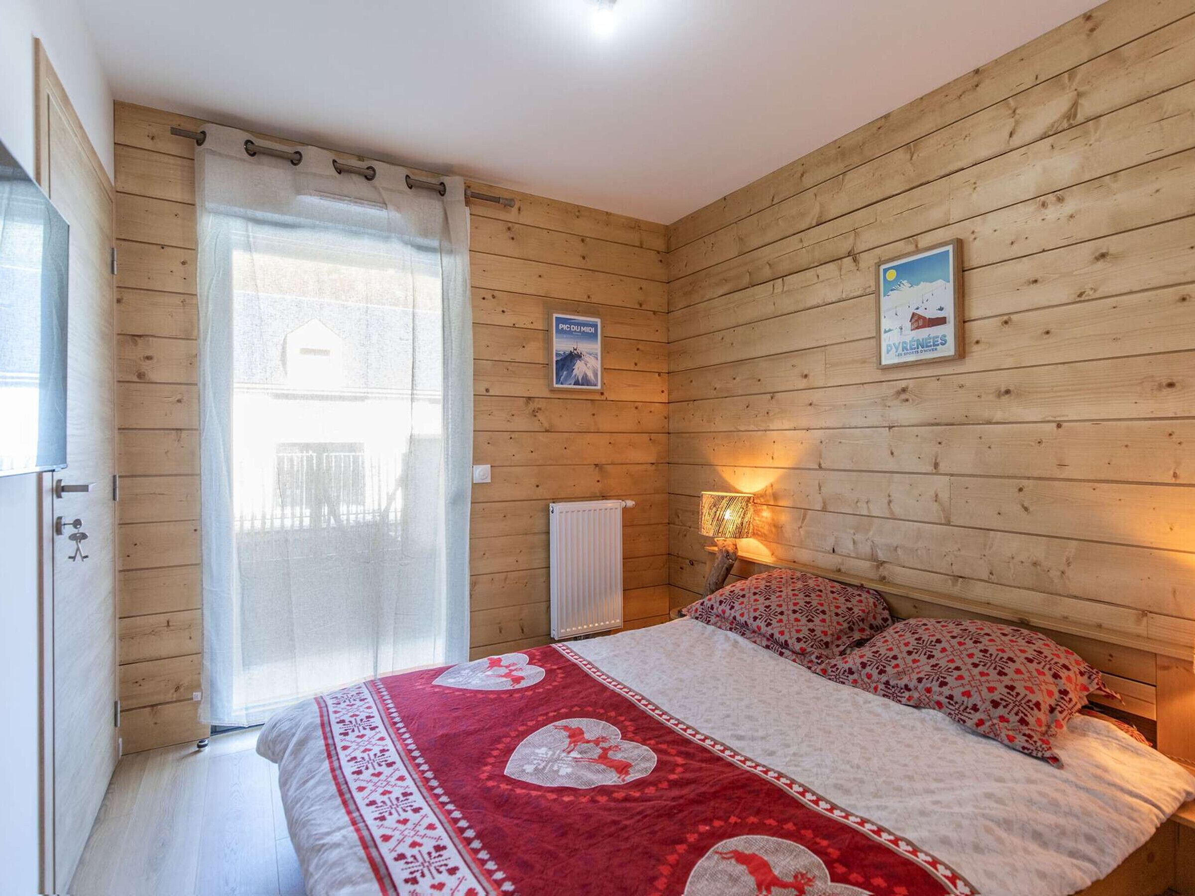 Une chambre confortable aux murs en bois, avec un lit recouvert d’une couette rouge et blanche, une fenêtre avec un rideau et une lampe fixée au mur.