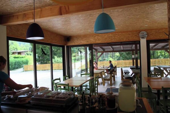 Un restaurant avec un plafond en bois, de grandes fenêtres, des tables et des chaises, et une personne qui travaille au comptoir.