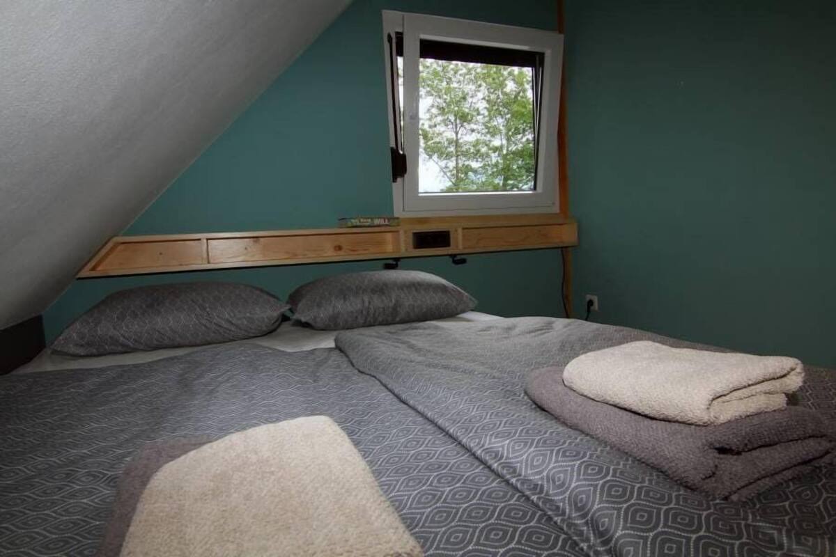 Ein Schlafzimmer mit schrägverdeckter Decke, einem Fenster und einem Bett mit gemusterten Betttextilien und Kissen.