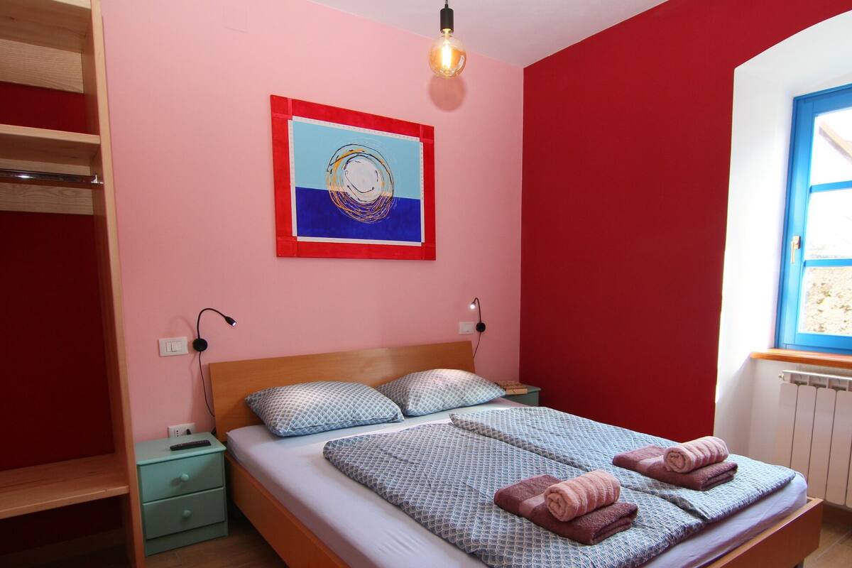 Ein Schlafzimmer mit einem Bett, zwei Kissen, einem Nachttisch, einer Wandlampe, einem Fenster und einem Gemälde.