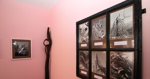 Een verzameling ingelijste kunstwerken aan een roze muur, waaronder een groot stuk in de vorm van een boom en kleinere ingelijste afbeeldingen.