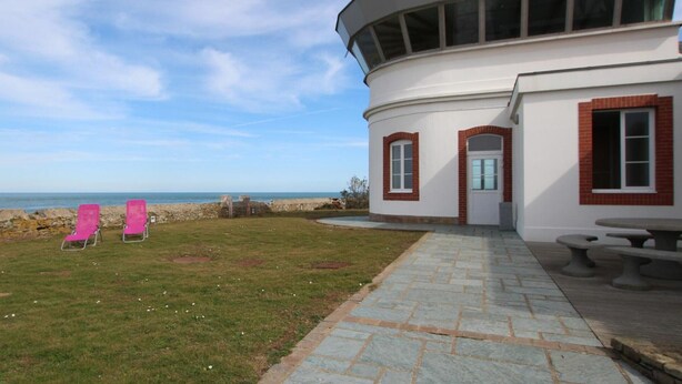 Un bâtiment situé sur la côte, doté d’un toit incurvé, d’une terrasse pavée et de deux fauteuils de jardin roses.
