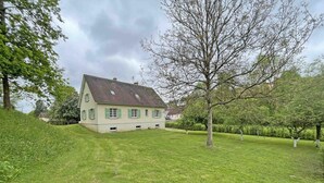 Uma casa grande com telhado inclinado, rodeada por um gramado bem cuidado e árvores.