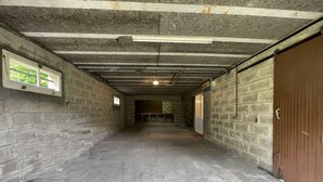 Um corredor com paredes de concreto, com pouca luz, e uma única janela à esquerda e uma porta fechada à direita.