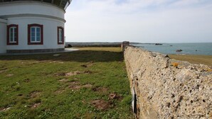 A coastal building with a round structure and a flat roof, adjacent to a grassy area and a stone wall overlooking the sea.