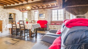 Una habitación acogedora con pared de piedra, vigas de madera, una mesa de comedor con sillas, un sofá con cojines rojos y una ventana con vistas.
