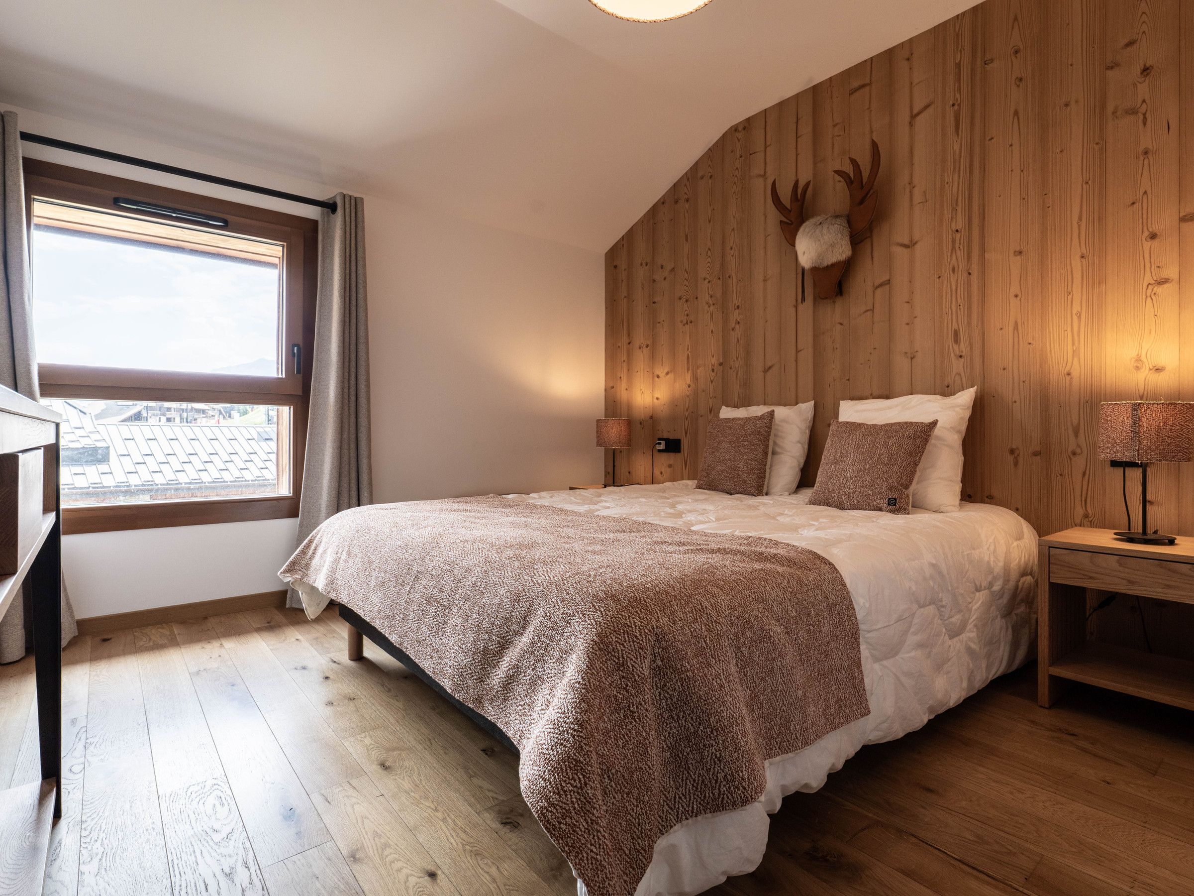 Une chambre à coucher comprenant un lit, une table de chevet, une lampe, une fenêtre et un mur en bois orné d’une tête de cerf.