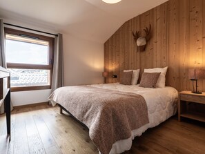 A bedroom with a bed, a nightstand, a lamp, a window, and a wooden wall with a mounted deer head. - Luxurious 3BR Apartment for 8, 50m from Slopes & Lift in Montalbert, WiFi, Parking (La Plagne Montalbert)