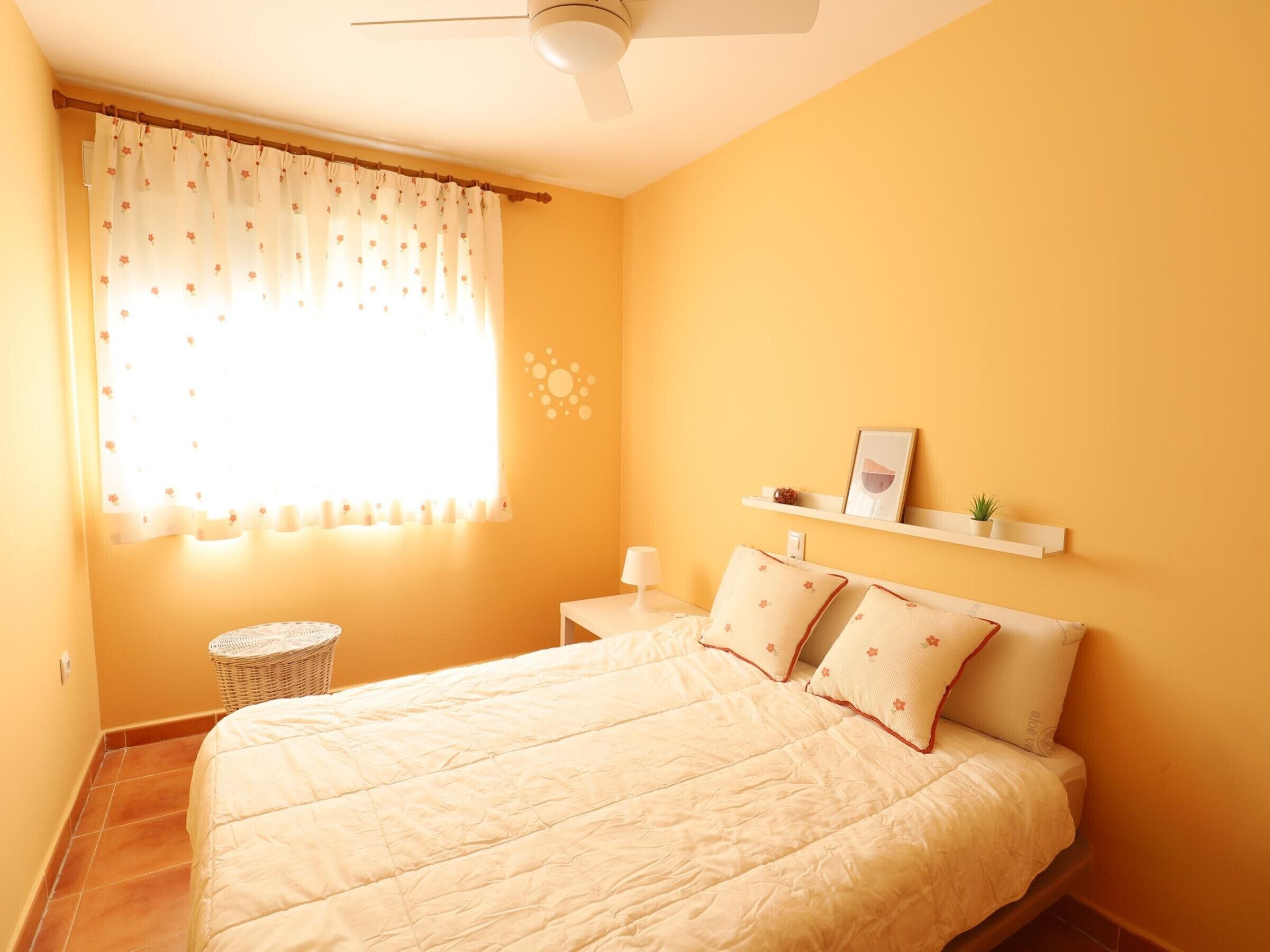 Un dormitorio con cama, mesita de noche, estante en la pared con objetos decorativos y ventana con cortinas florales.