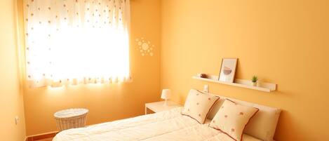 Un dormitorio con cama, mesita de noche, estante en la pared con objetos decorativos y ventana con cortinas florales.