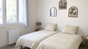 Een slaapkamer met twee bedden, een radiator, een raam met gordijnen, een nachtkastje met lamp en wanddecoratie.
