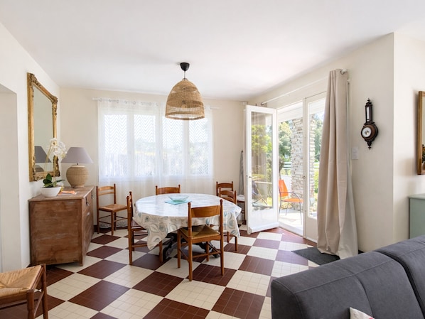 A dining area with a checkered floor, a round table, and chairs. There is a window with curtains, a hanging light fixture, and a clock on the wall.