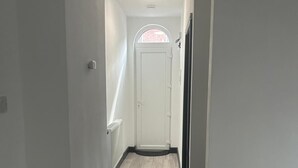 Un pasillo estrecho con una puerta blanca al final, una ventana pequeña y una lámpara en el techo.