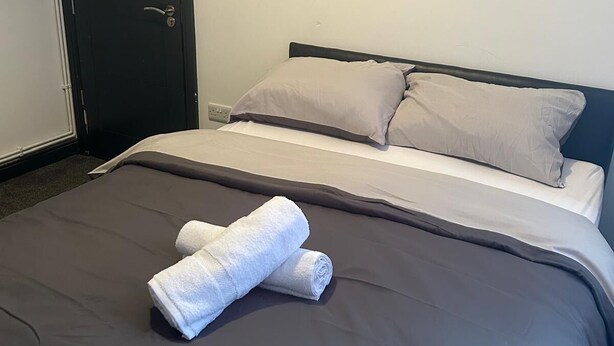 Una cama bien hecha con ropa de cama gris y dos toallas blancas enrolladas, una puerta oscura y una pared blanca lisa.